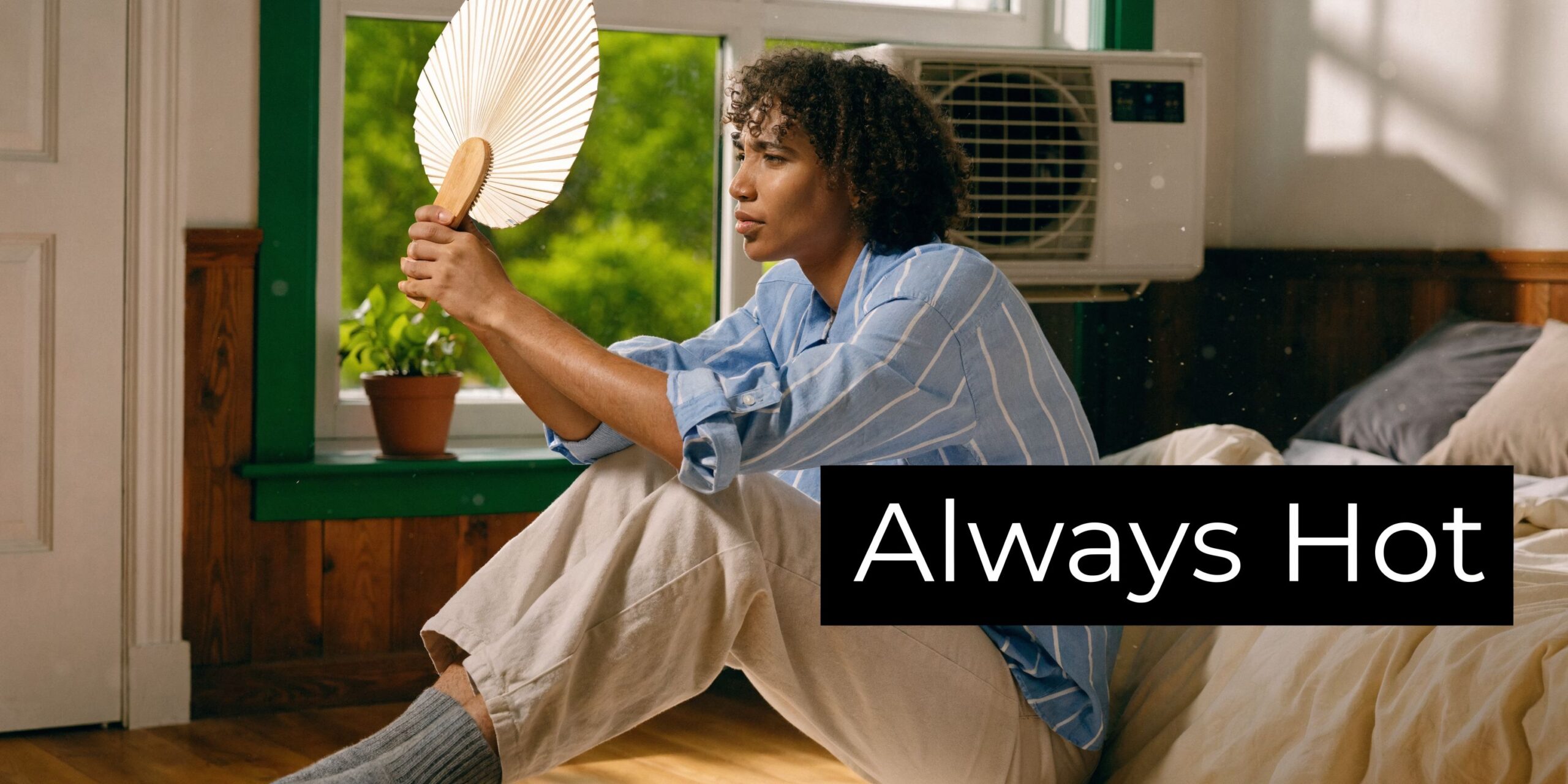 A person sitting on a bed fanning themselves with a hand fan near an air conditioner unit.